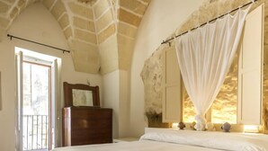 A bedroom with a stone ceiling, a wooden dresser, a bed with white bedding, and a window with a white curtain.