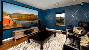 Ein modernes Wohnzimmer mit einem großen Flachbildfernseher, einem schwarzen Ledersofa, einem Holztisch und einer blauen Wand mit geometrischen Mustern.