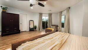 Ein Schlafzimmer mit einem Bett, einem Deckenventilator, einem Kleiderschrank, einem großen Spiegel und Blick ins Freie durch ein Fenster.