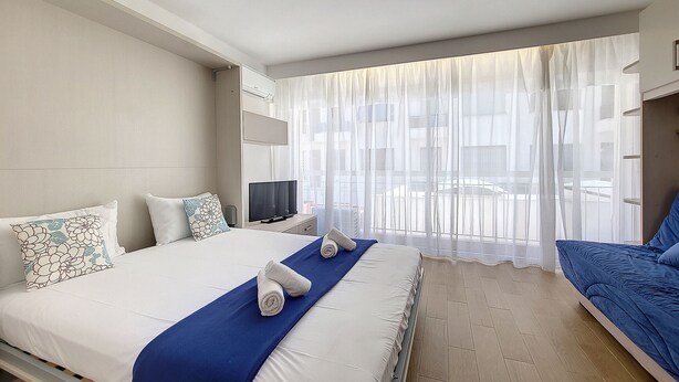 A hotel room with a bed, a TV, and a window with sheer curtains.