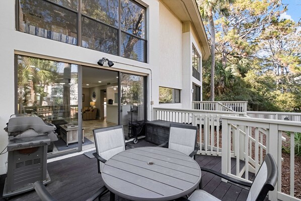 An outdoor patio with a round table and chairs, a grill, and a view of the house's interior through large glass doors.