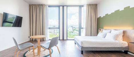 A modern hotel room with a bed, a desk with two chairs, a round table, and a large window with curtains.