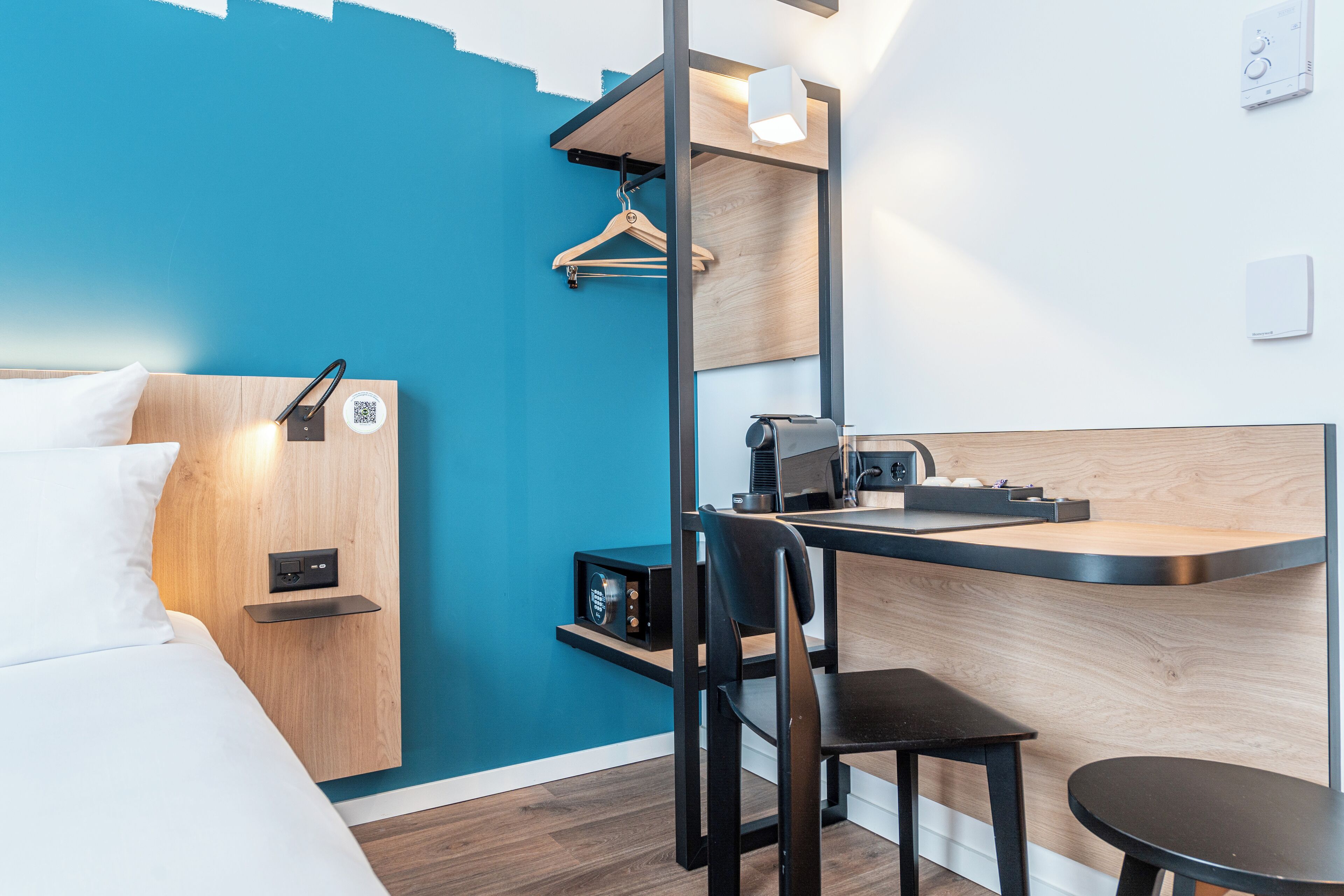 A modern hotel room with a bed, a desk, a chair, and a small kitchenette.