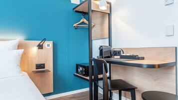 A modern hotel room with a bed, a desk, a chair, and a small kitchenette.