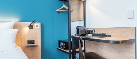 A modern hotel room with a bed, a desk, a chair, and a small kitchenette.