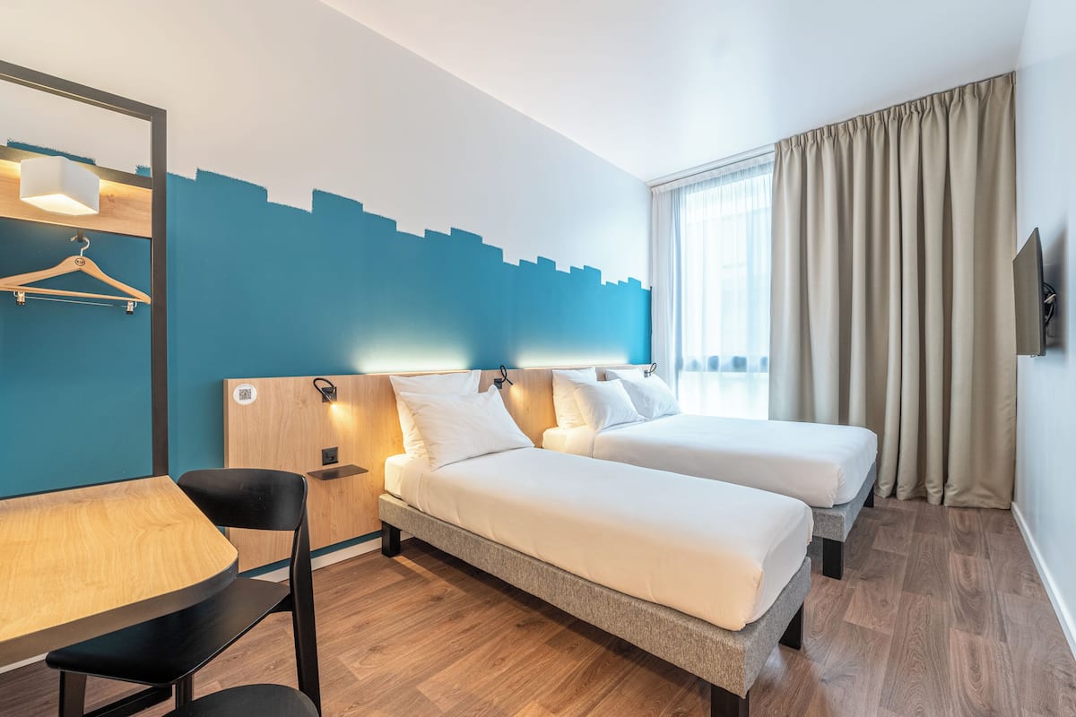 Hotellihuone, jossa on kaksi sänkyä, työpöytä, tuoli, puinen pöytä, riippuvalaisin ja suuri ikkuna, jossa on verhot.