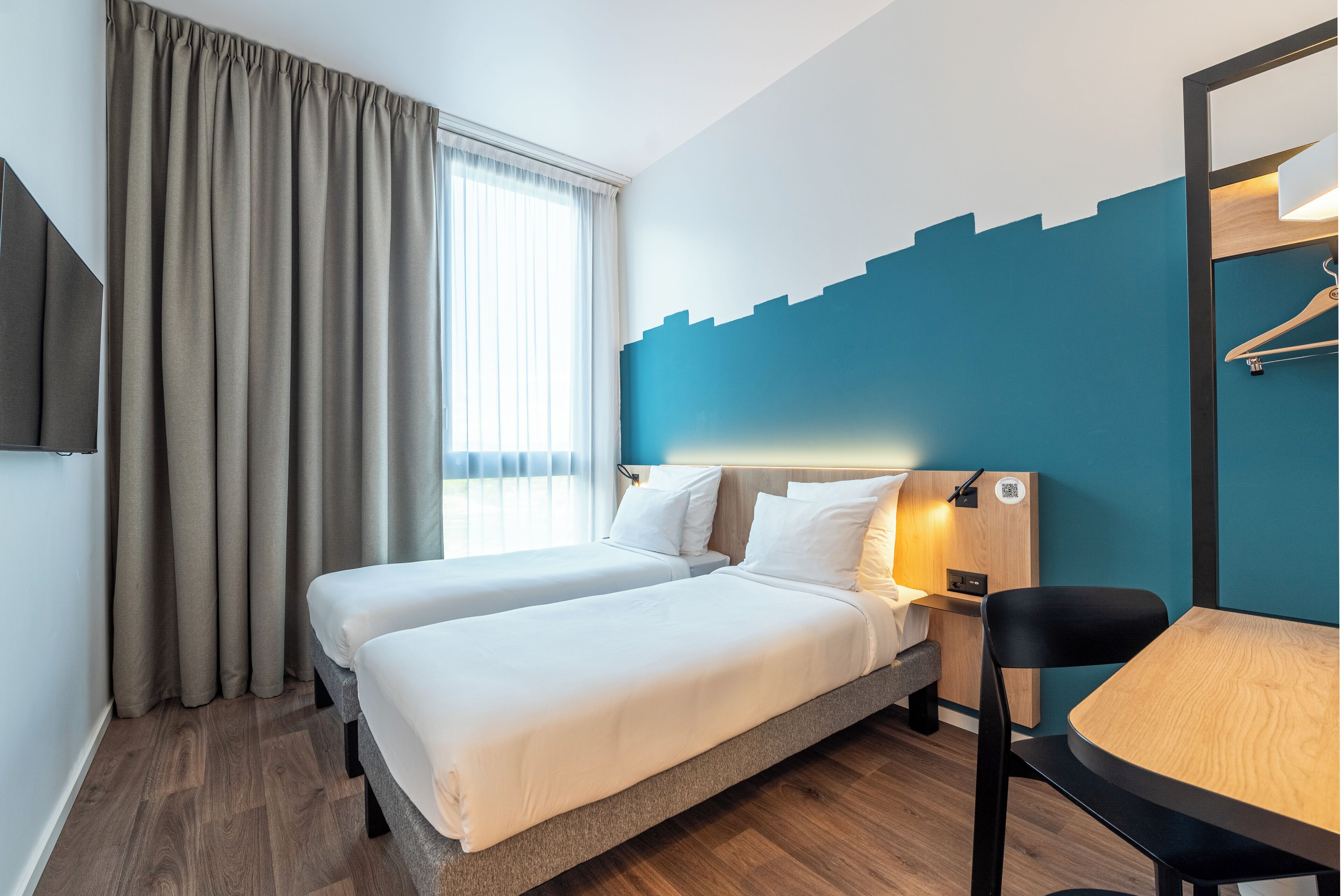 A hotel room with two beds, a desk, a chair, a TV, and a window with curtains.