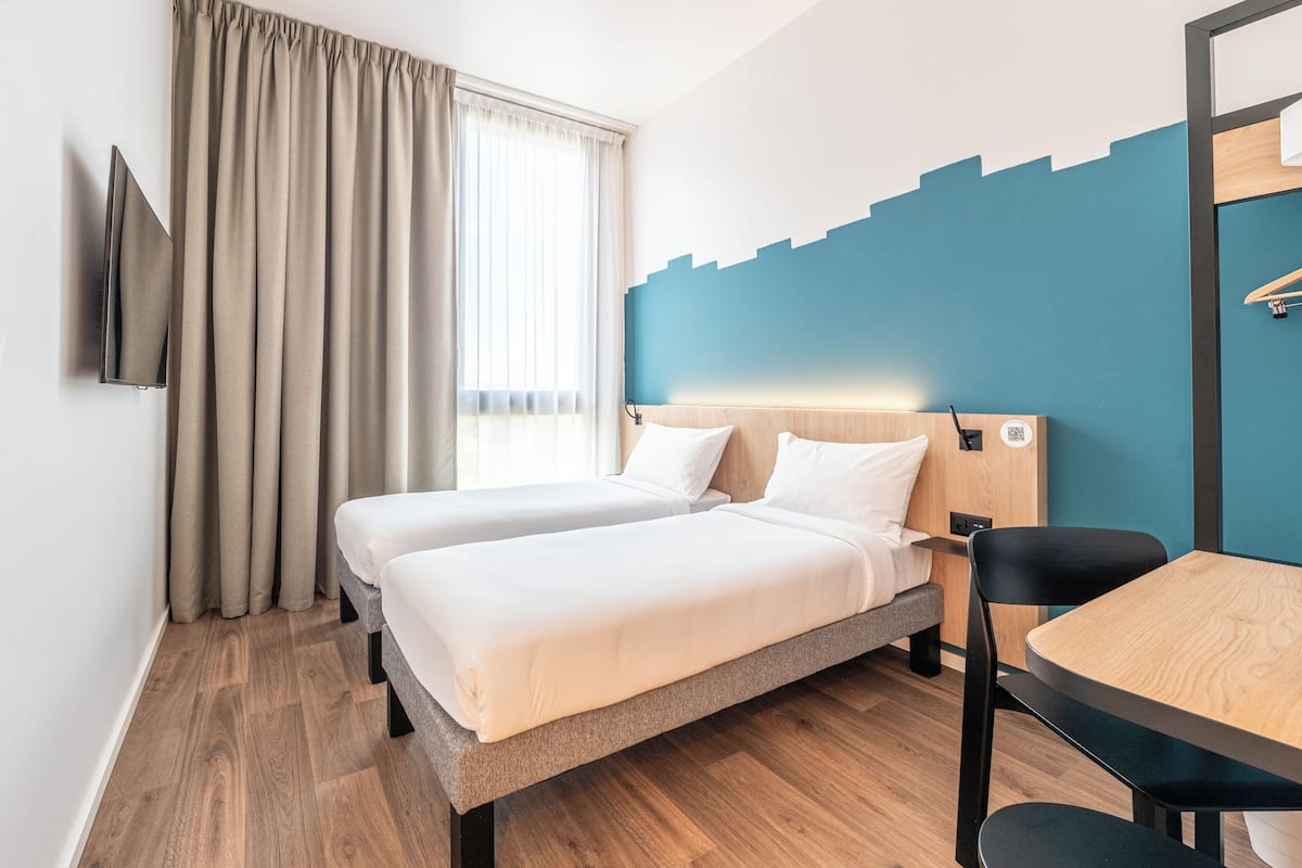 Hotellihuone, jossa on kaksi sänkyä, puinen pöytä, tuoli, seinälle asennettu televisio ja ikkuna, jossa on verhot.