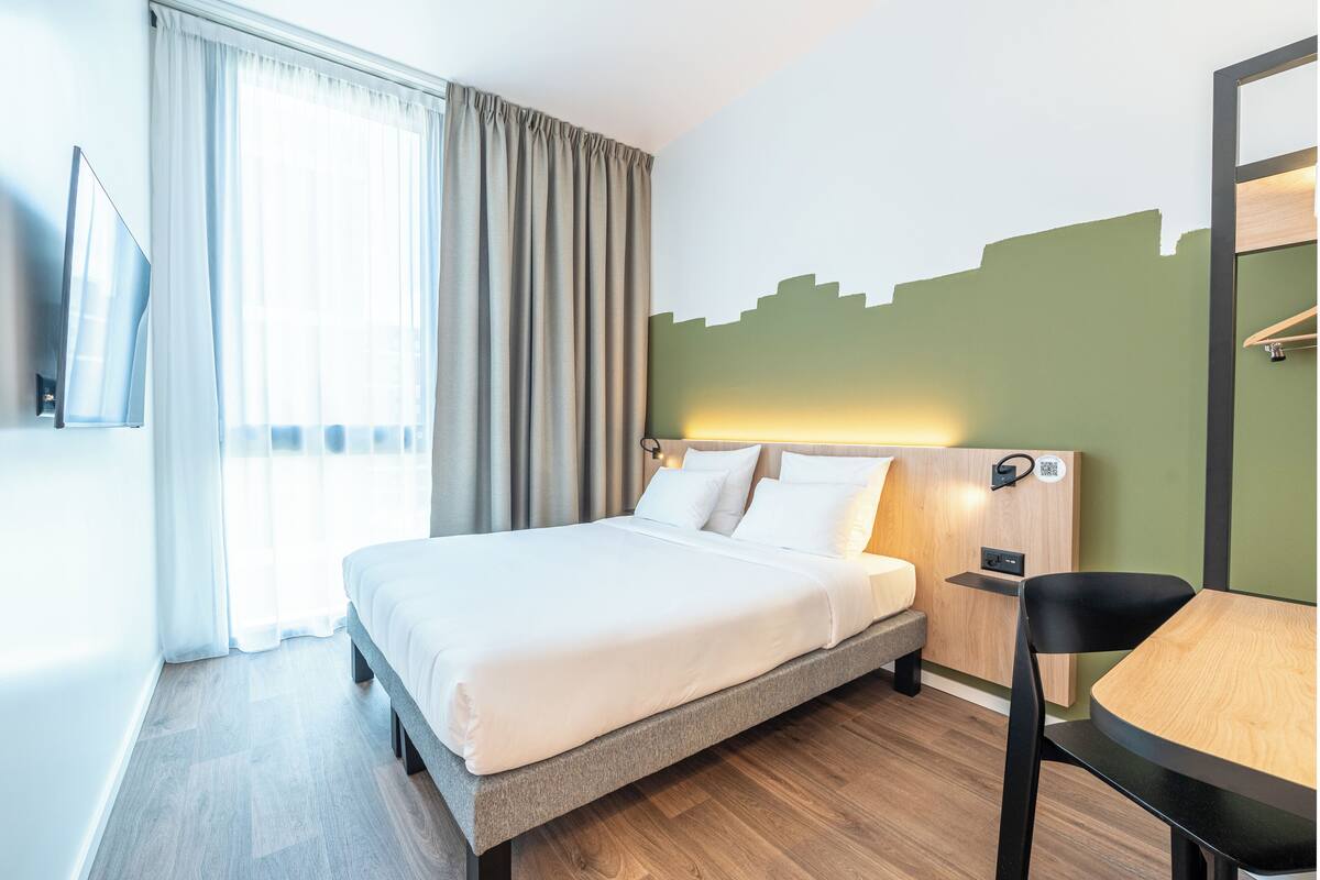 Hotellihuone, jossa on sänky, työpöytä, tuoli, televisio ja ikkuna, jossa on verhot.