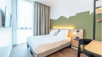 Quarto de hotel com cama, escrivaninha, cadeira, televisão e janela com cortinas.