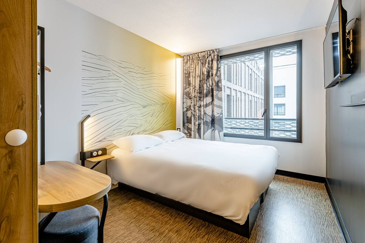 Hotelkamer met een bed, bureau, stoel, televisie, raam met uitzicht en een kunstwerk aan de muur.