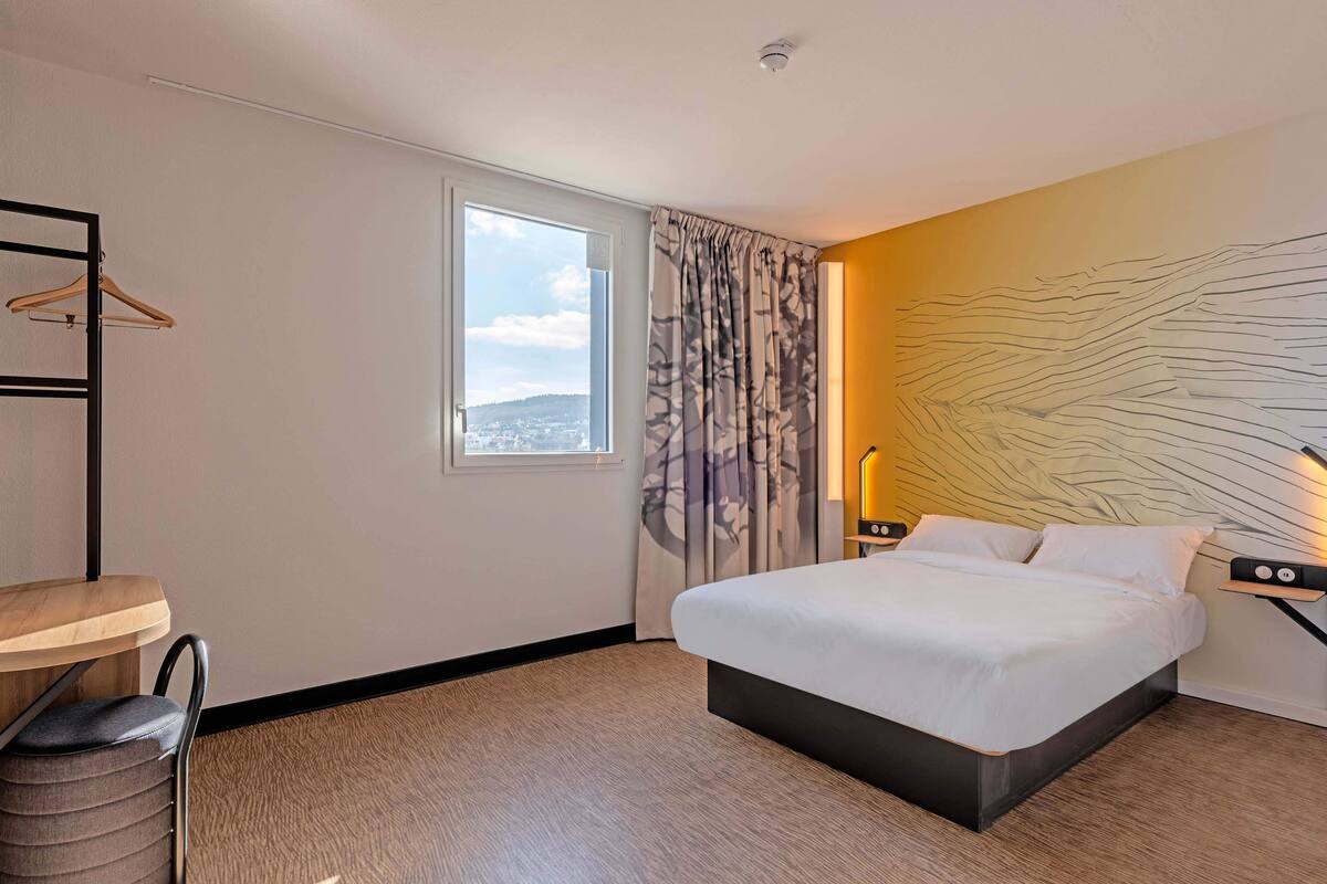 Een hotelkamer met een bed, een raam, een hanglamp en een kunstwerk aan de muur.