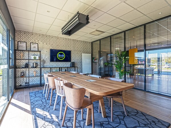 A modern conference room with a wooden table, beige chairs, a TV screen displaying 'B&B HOTELS', and a large window with blinds.