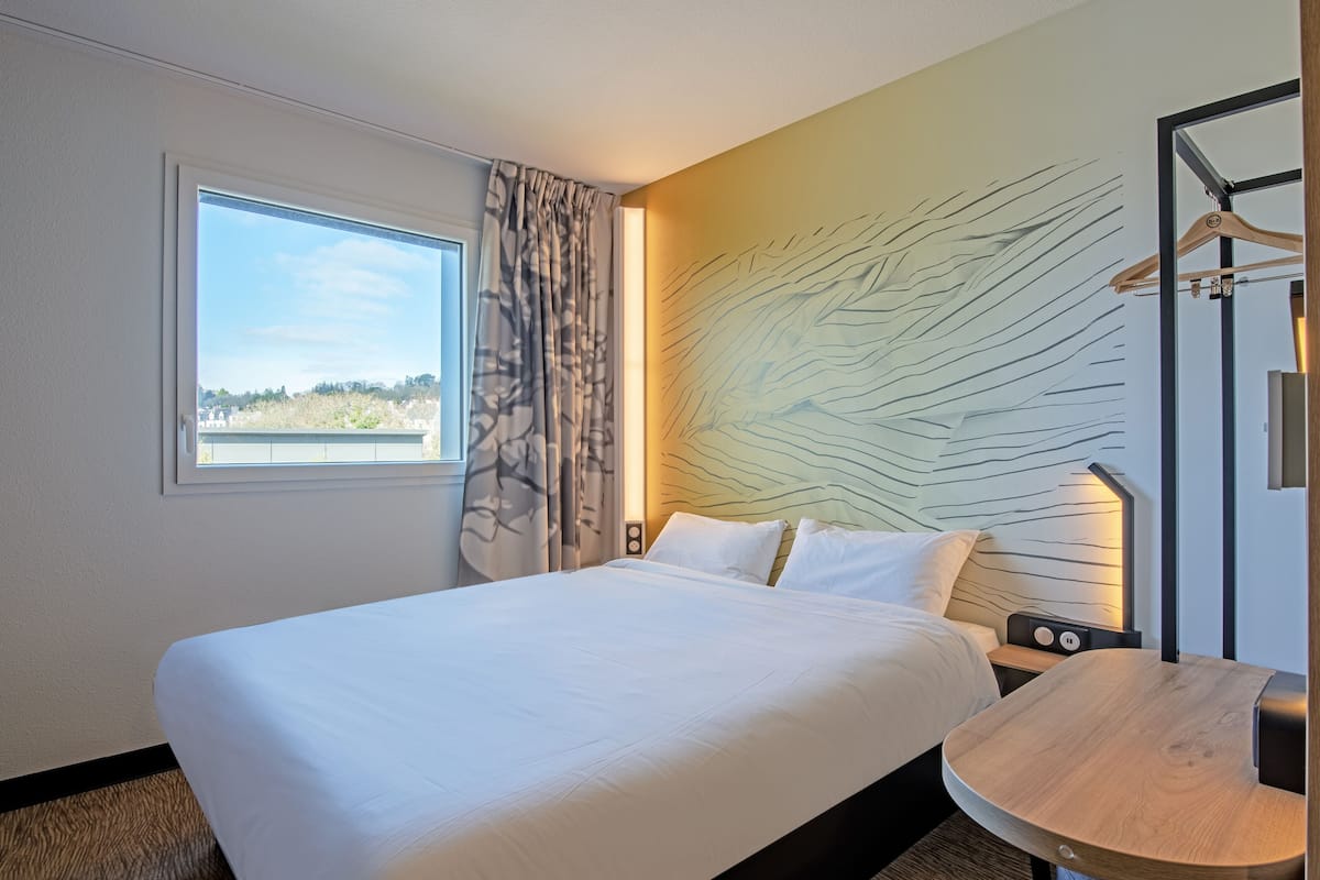 Een hotelkamer met een bed, een klein tafeltje, een aan de muur bevestigde televisie en een raam met gordijnen.