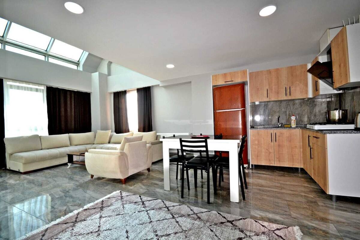 A modern living room with a sofa, dining table, and chairs, a kitchen with wooden cabinets, and a large window providing natural light.
