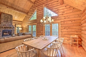 L’intérieur d’une cabane en rondins, avec une cheminée en pierre, une table à manger en bois, des chaises blanches et une grande fenêtre.