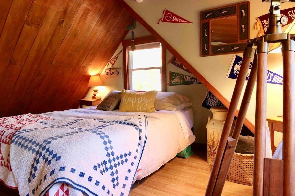 A cozy attic bedroom with a wooden sloped ceiling, a bed with a quilt, a lamp, and a telescope on a tripod.