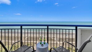 A balcony with a view of a beach and ocean. There are two chairs and a round table with two bottles of beer.
