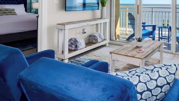 A beachfront property with a balcony, a bed, blue furniture, and a television.