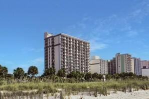 Un hotel moderno de gran altura con varios balcones, rodeado por una playa de arena y palmeras.