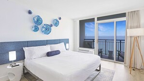 A hotel room with a large bed, a view of the sea through a balcony, and decorative blue glass art on the wall.