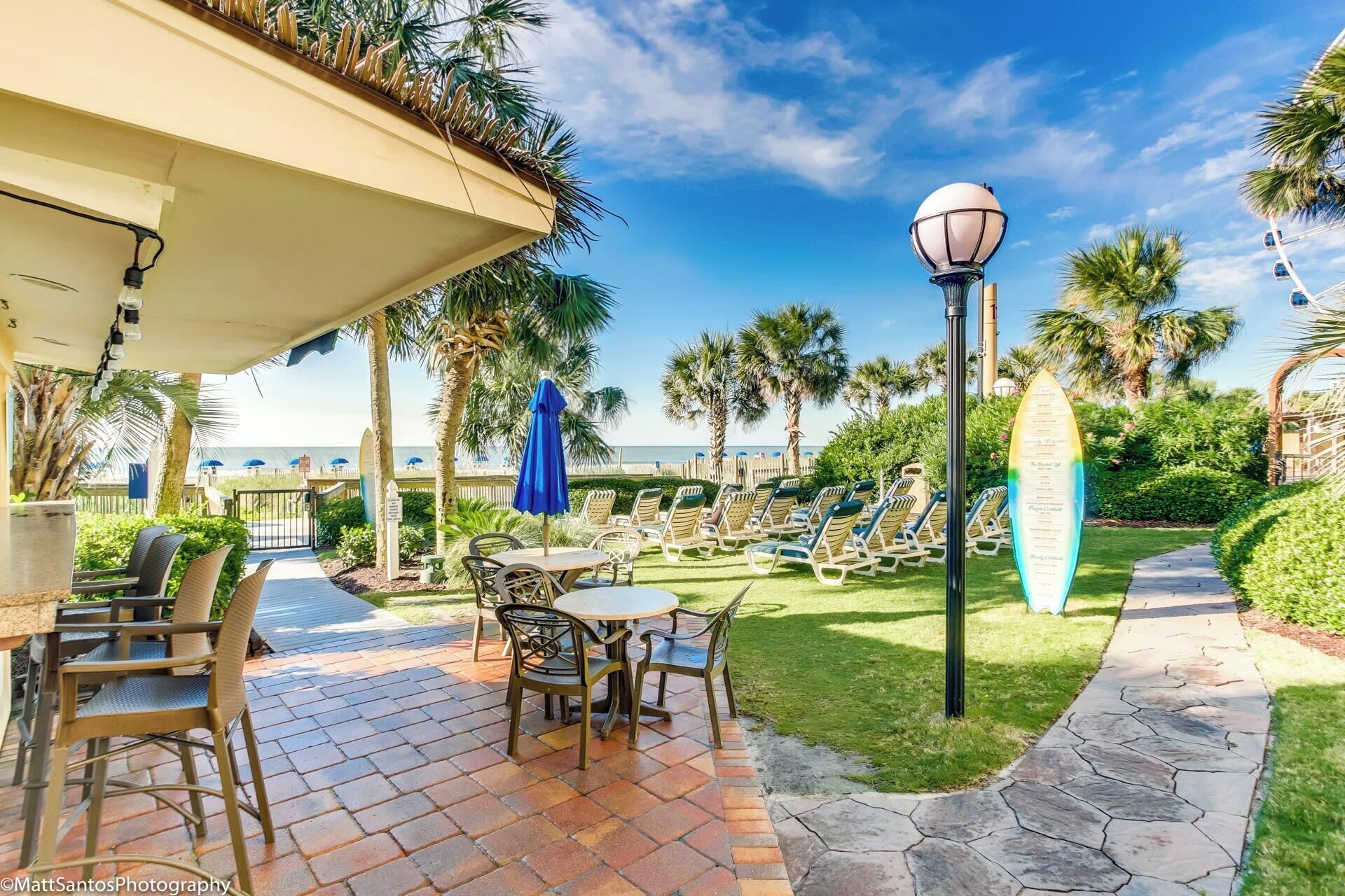 A patio area with outdoor seating, a surfboard-shaped sign, and a view of the ocean.