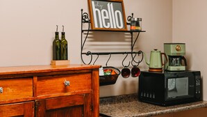 A kitchen with a wooden cabinet, a wall-mounted shelf with a 'hello' sign, a green coffee maker, a black microwave, and two olive oil bottles.