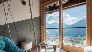 Ein Zimmer mit großem Fenster, einem Hängekorbstuhl, einem kleinen Tisch mit Buch und Tasse sowie einem flauschigen weißen Kissen.