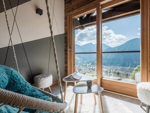 A room with a large window, a hanging chair, a small table with a book and mug, and a fluffy white cushion.