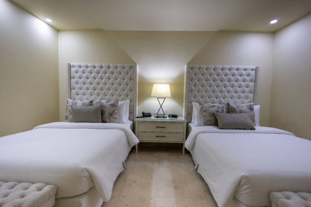 Une chambre d’hôtel avec deux lits, chacun doté d’une tête de lit capitonnée, d’une table de chevet avec une lampe et d’un petit tiroir.