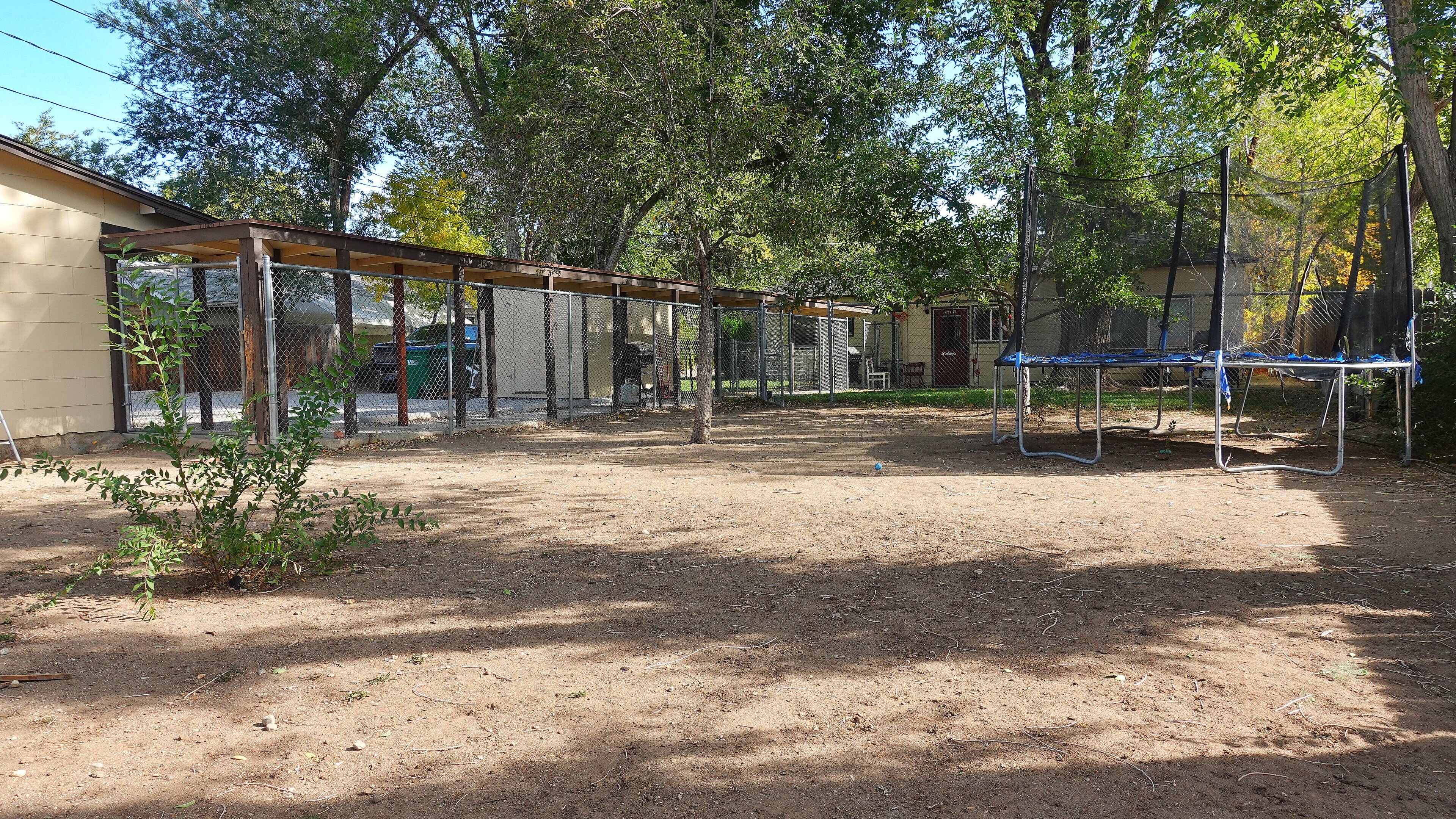 A fenced outdoor area with a trampoline, trees, and a building in the background.