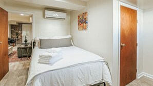 Un dormitorio con cama, unidad de aire acondicionado, puerta de madera y una obra de arte colorida en la pared.