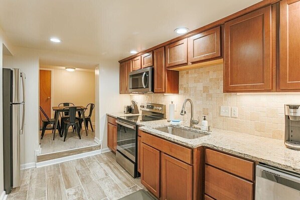A modern kitchen with wooden cabinets, a granite countertop, stainless steel appliances, and a dining area with a table and chairs. - Staunton Underground: Center of Downtown (Staunton)