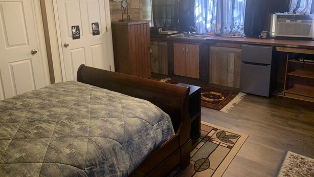 A bedroom with a bed, a wooden bench, a small kitchenette with a refrigerator, and a window with curtains. - Cozy Studio apartment with cabin feel (Tallahassee)