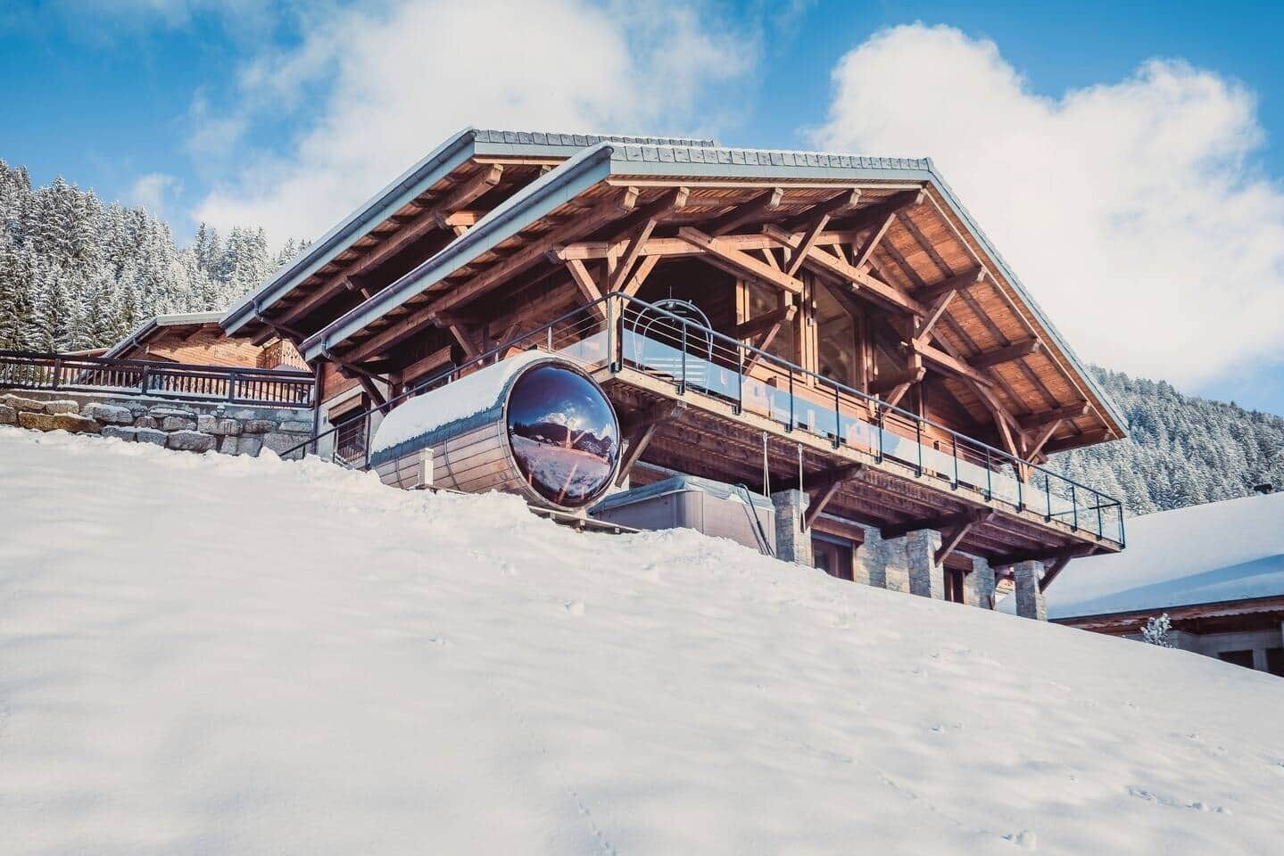 A wooden chalet with a balcony, set against a snowy mountain backdrop.
