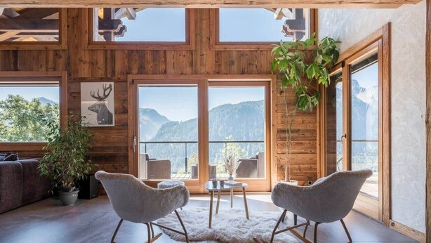Een ruime kamer met een groot raam, houten balken in het plafond, een zacht, wit vloerkleed en twee schommelstoelen.