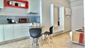 Cocina moderna con microondas, cafetera y hervidor sobre la encimera. Hay una mesa de comedor con sillas y una cama con manta.