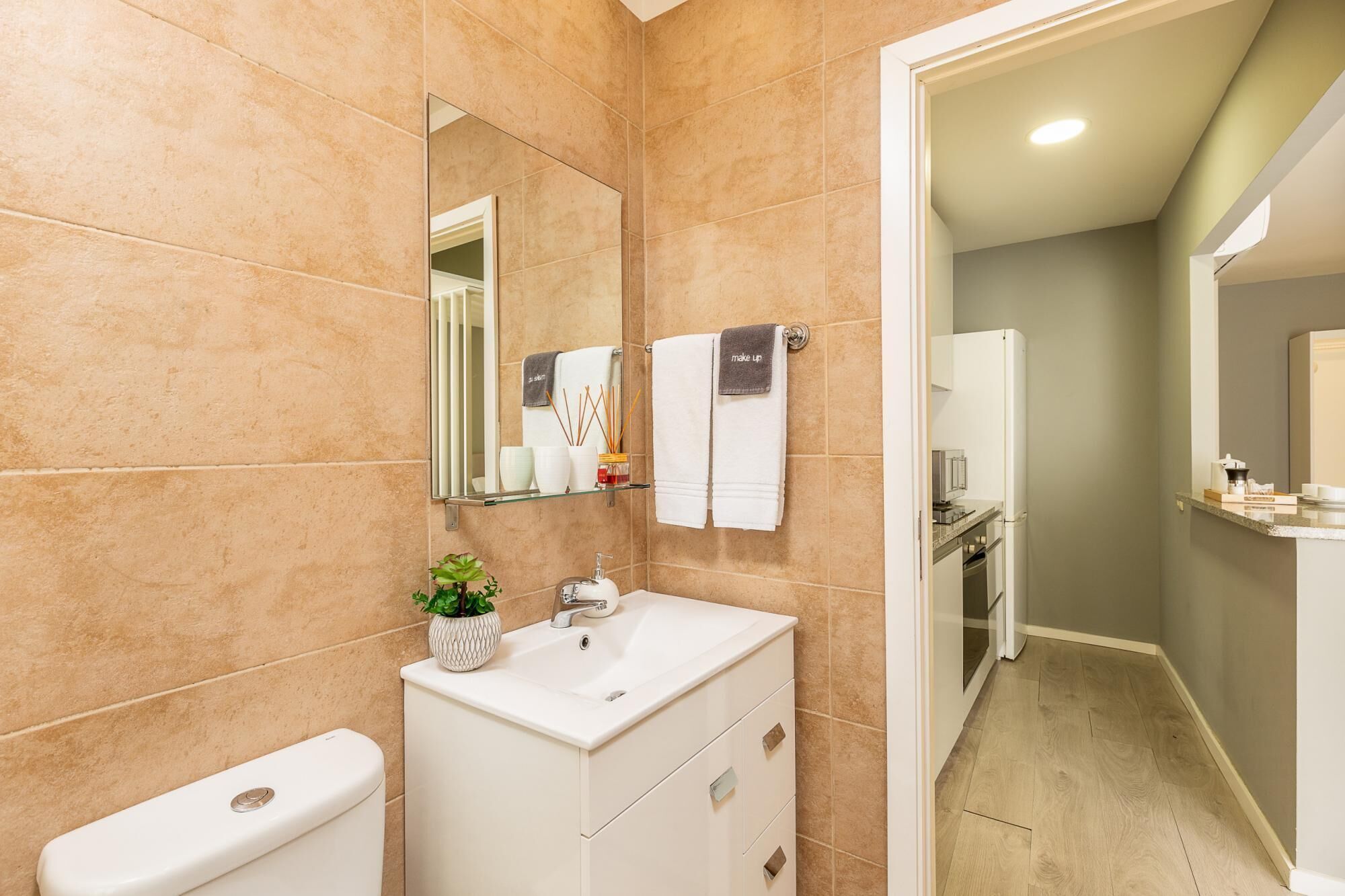 A modern bathroom with beige tiles, a white sink, a mirror, and a toilet. There is a small plant on the sink and a towel rack with two towels.