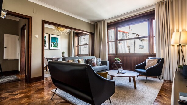 Een moderne woonkamer met een bank, fauteuil en salontafel, grote ramen met gordijnen en uitzicht op gebouwen.
