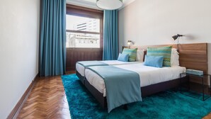 Een moderne slaapkamer met twee bedden, een groot raam met gordijnen, een houten hoofdbord en een blauwgroen vloerkleed.