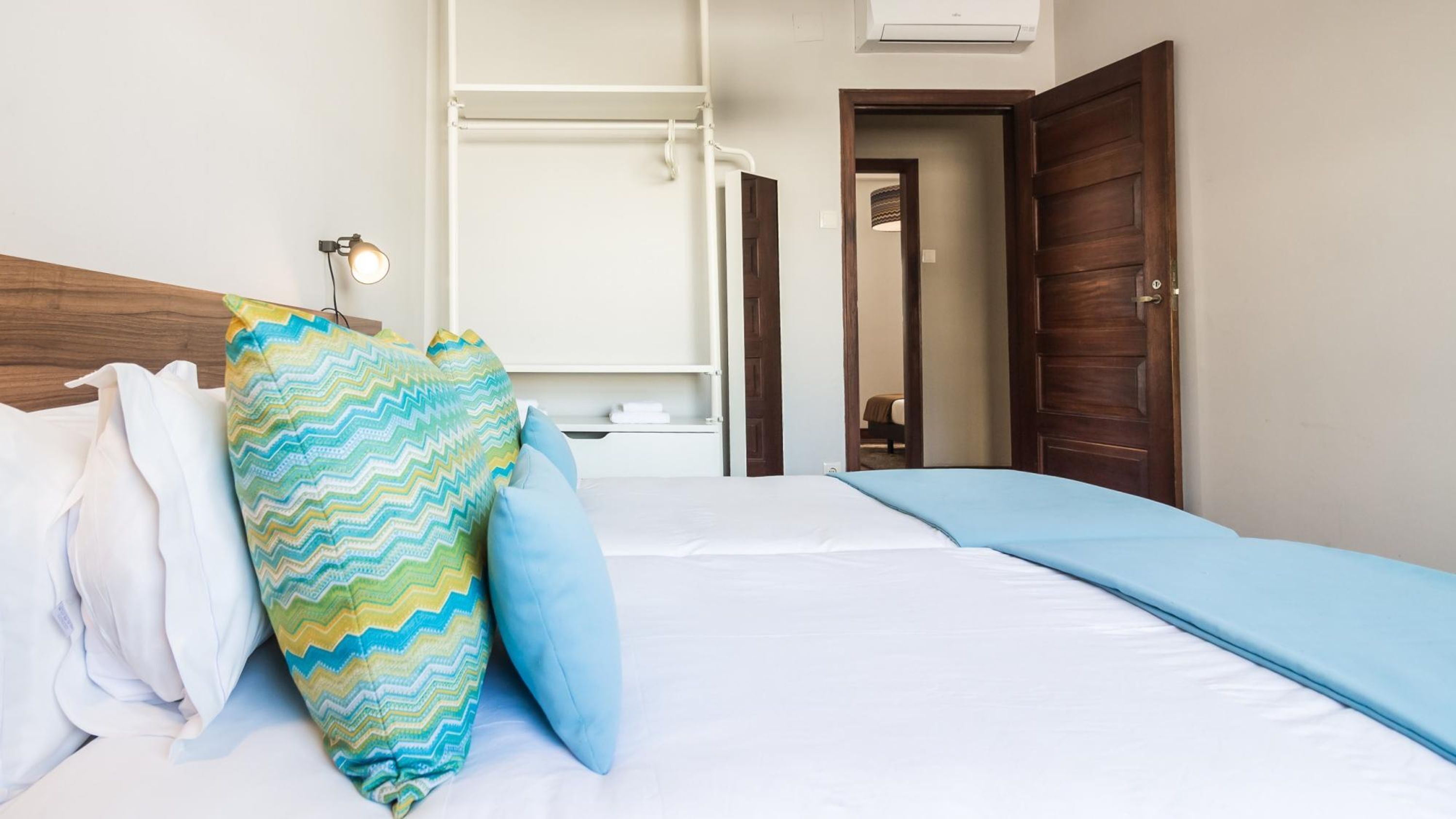 A hotel room with a bed, a bedside lamp, a wooden headboard, and a door leading to another room.