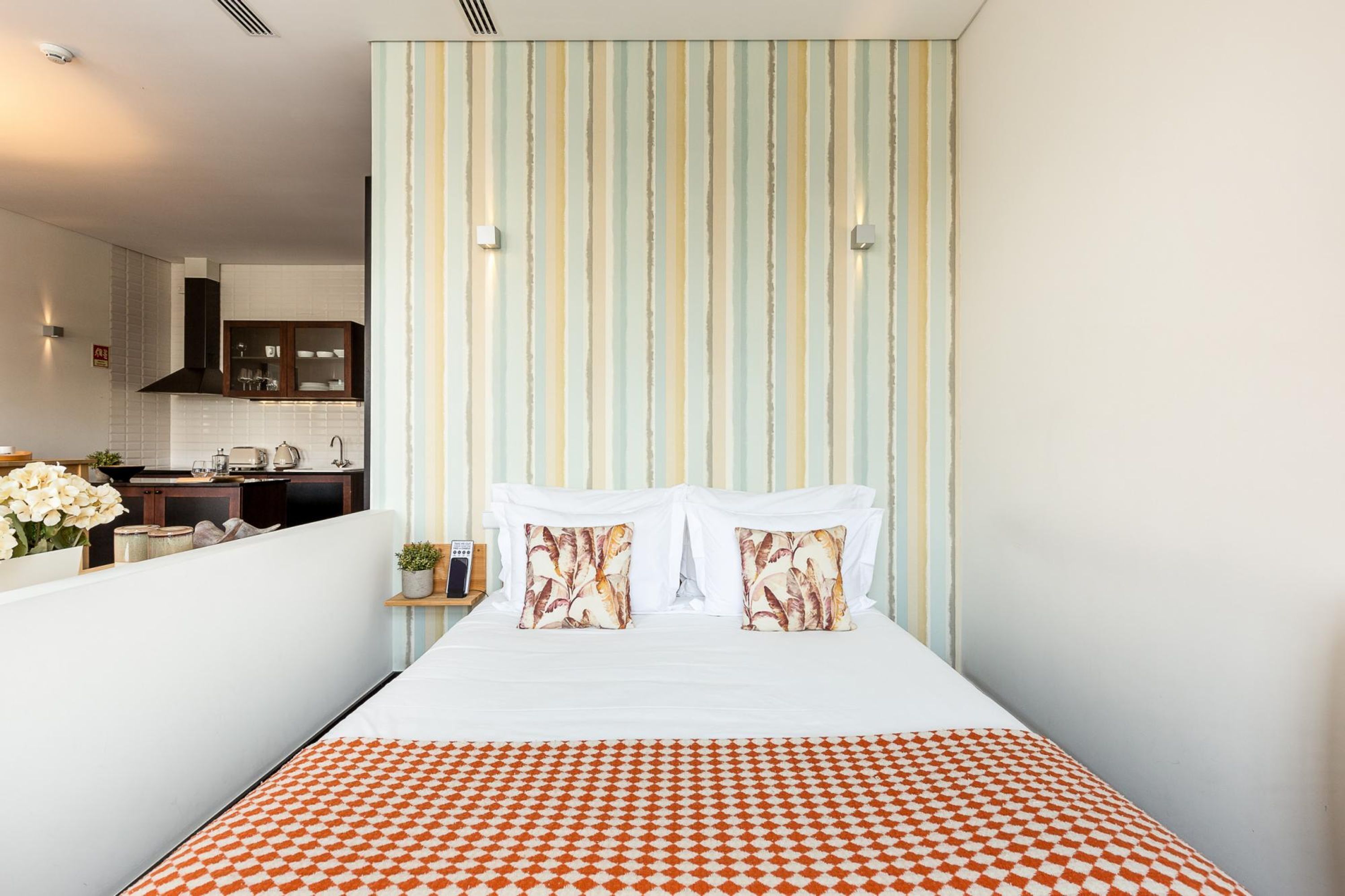 Habitación de hotel estrecha con una cama, papel tapiz a rayas y una cocina visible a través de una puerta abierta.