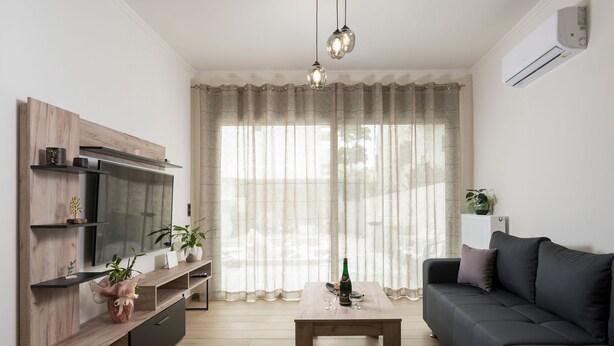 A modern living room with a flat-screen TV, wooden shelving, a coffee table, a sofa, and a large window with sheer curtains. - Villa Cataleya : A Spacious, Private Oasis with a Pool Near the Beach (Chania)
