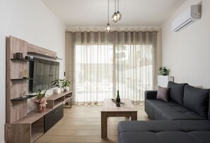 Ein modernes Wohnzimmer mit Flachbildfernseher, Holzregalen, einem Couchtisch, einer Couch und einem großen Fenster mit durchsichtigen Vorhängen.