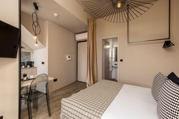 A modern hotel room with a bed, a desk with a chair, a bathroom, and a ceiling light fixture.