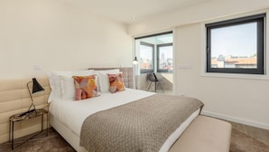 A bedroom with a bed, a bedside table with a lamp, a chair, and a large window offering a city view. - RA - Santa Catarina in Oporto (Porto)