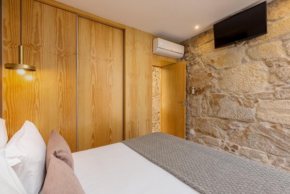 A bedroom with a bed, wooden wardrobe, stone wall, and a mounted TV.