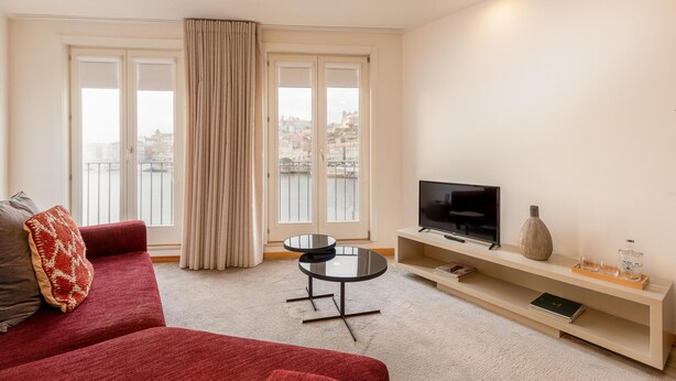 A modern living room with a red sofa, a small round table, a TV on a stand, and a view of buildings through large windows. - Ribeira Gaia 3 in Vila Nova de Gaia (Vila Nova de Gaia)