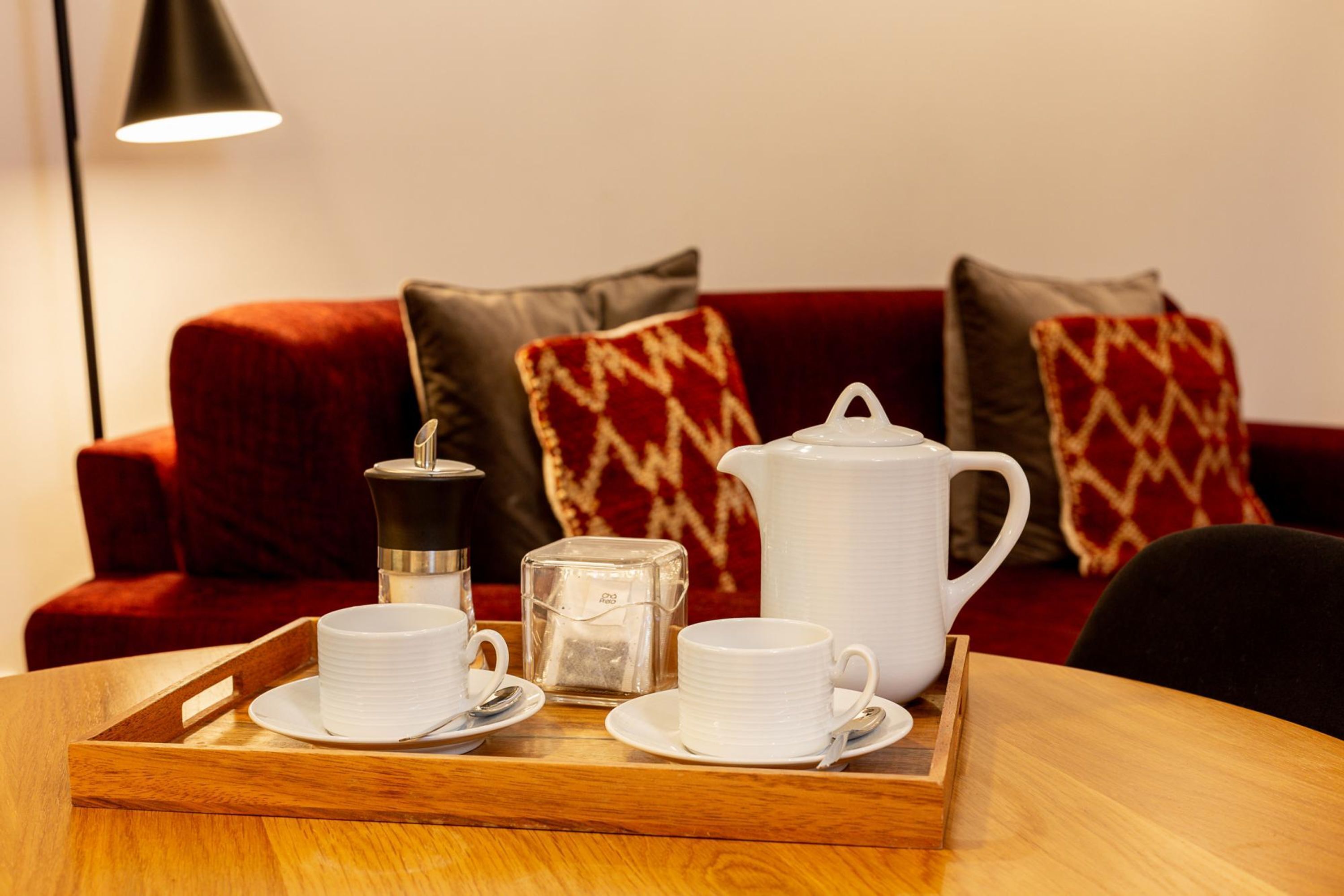 A wooden tray with white cups, a teapot, and a sugar container on a table in front of a red sofa with patterned cushions.