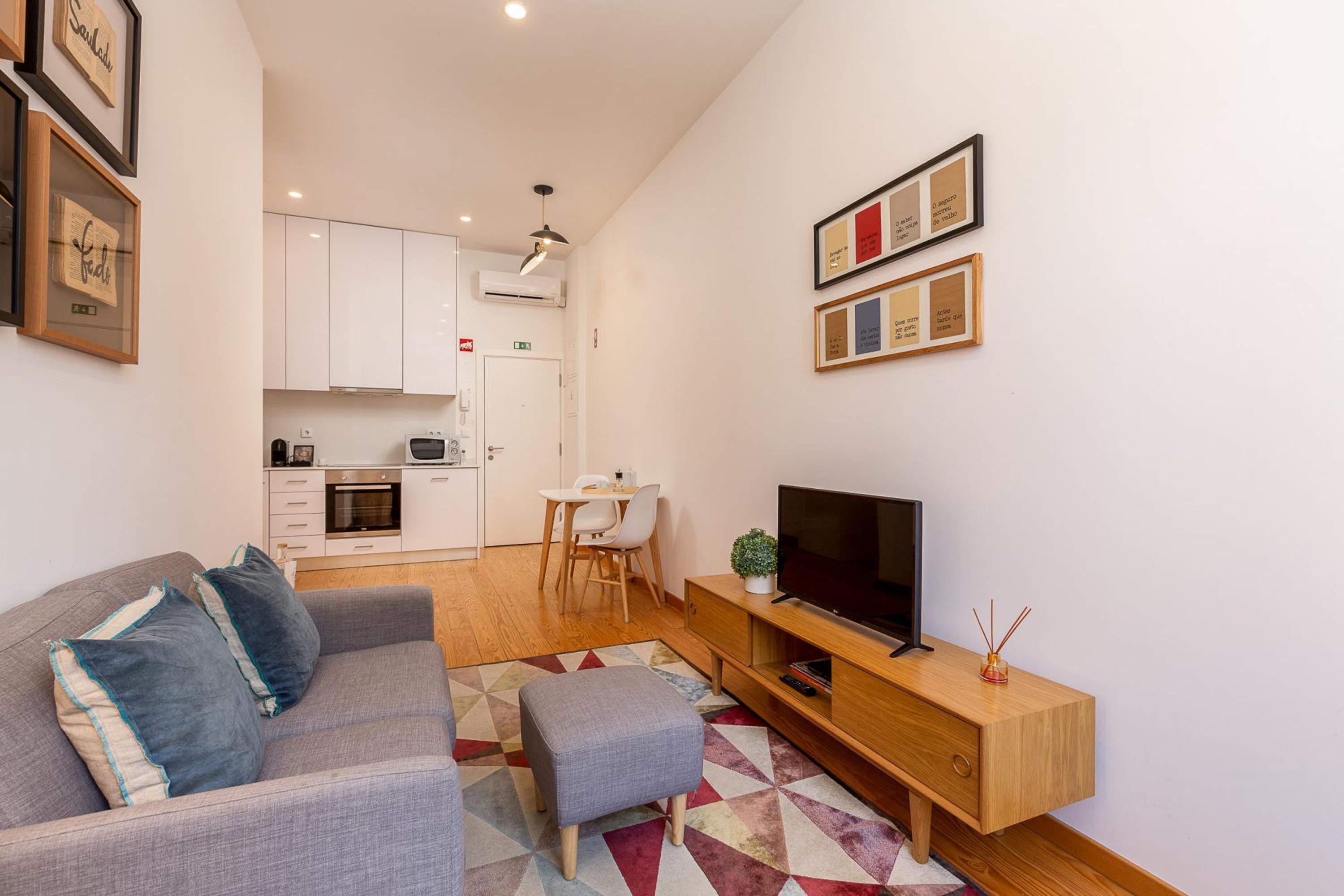 Uma sala de estar moderna com um sofá cinzento, uma televisão e um móvel para TV de madeira. Ao fundo, cozinha com armários brancos e um espaço de jantar com mesa e cadeiras.